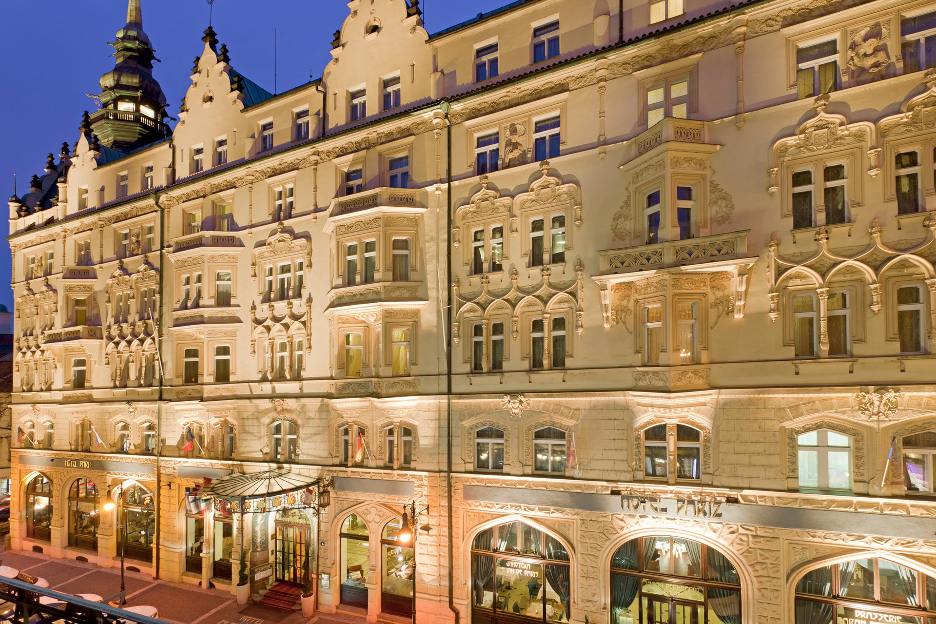 The Hotel Paris Prague lit up at dusk, its Neo-Gothic and Art Nouveau façade and corner tower above the street